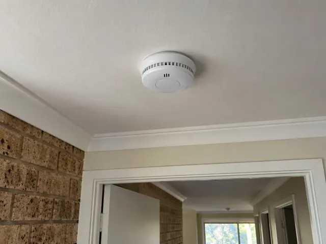 Ceiling-mounted smoke alarm in a cream hallway with brick walls and white trim in Maudsland